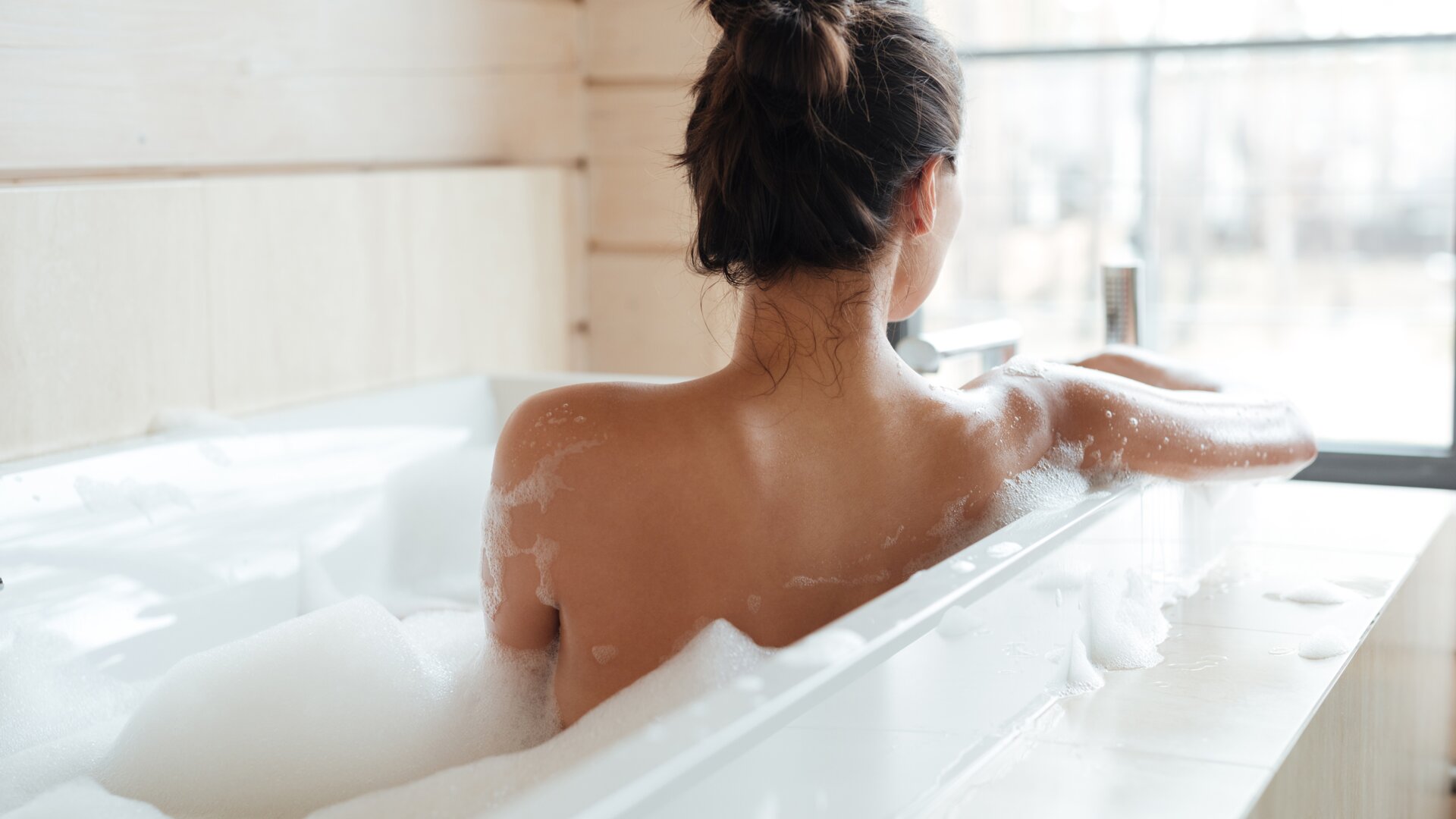 Achterkant van een vrouw zittend in een witte badkuip gevuld met schuim die uit het raam naar de natuur kijkt.